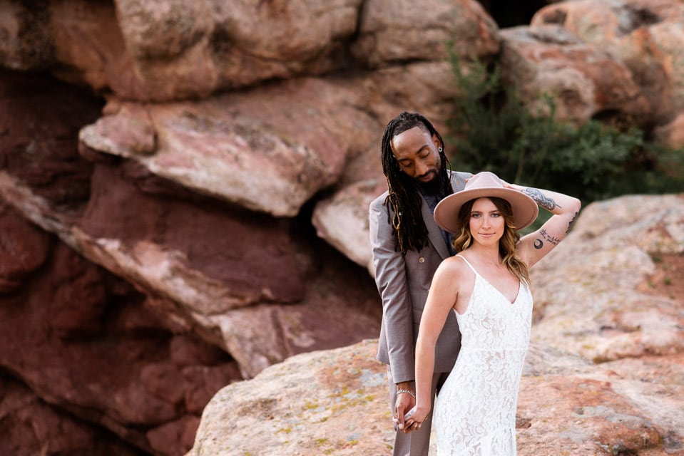 Bohemian Colorado elopement at Red Rocks. Boho elopement inspiration among the red rock formations near Morrison, Colorado. Groom stands behind bride while she holds onto her hat looking at the camera.